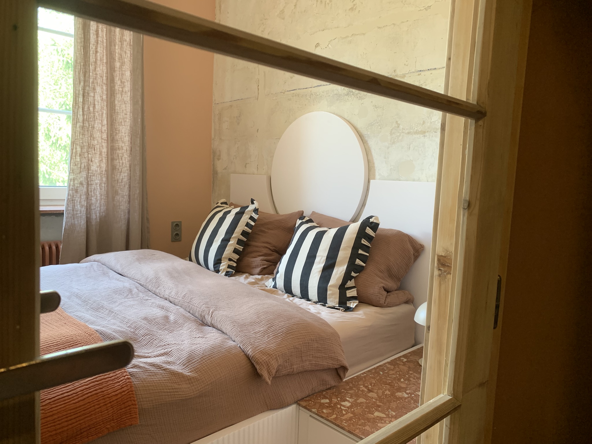 Kleines Schlafzimmer 36 qm mit Marmor-Wand, großem rundem weißem Spiegel, zwei gestreiften Baumwoll-Kissen, cremefarbenem Bett, weißem Stauraum-Schrank mit offenen Regalen, Licht durch Leinenvorhänge von links