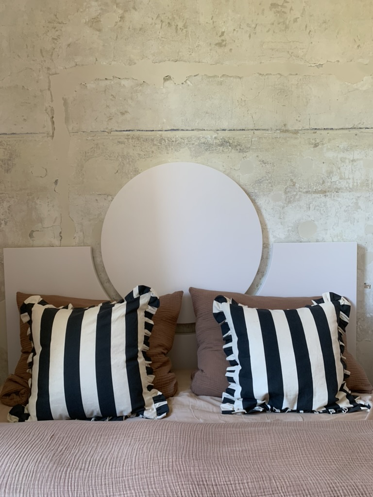 Blick durchs Türglas auf ein Schlafzimmer in Wien, 36 Quadratmeter. Das Bett hat ein Kopfteil aus weißem Marmor, dahinter eine Wand aus hellem Travertin mit sichtbaren Fugen. Zwei Kissen mit schwarzen und weißen Streifen und Rüschenrand liegen auf der Bettwäsche. Daneben ein drittes Kissen in Braun. An der Ecke neben dem Bett steht ein weißer MDF-Schrank mit geschlossenen Türen und einem offenen Regal mit drei Böden. Der Spiegel hinter dem Bett ist rund und matt. Ein dünner Leinenvorhang fällt vom Fenster. Der Boden ist Terrakotta, warm und matt.