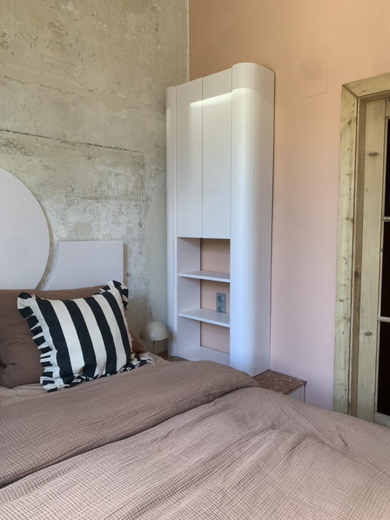 Blick durchs Türglas auf ein Schlafzimmer in Wien, 36 Quadratmeter. Das Bett hat ein Kopfteil aus weißem Marmor, dahinter eine Wand aus hellem Travertin mit sichtbaren Fugen. Zwei Kissen mit schwarzen und weißen Streifen und Rüschenrand liegen auf der Bettwäsche. Daneben ein drittes Kissen in Braun. An der Ecke neben dem Bett steht ein weißer MDF-Schrank mit geschlossenen Türen und einem offenen Regal mit drei Böden. Der Spiegel hinter dem Bett ist rund und matt. Ein dünner Leinenvorhang fällt vom Fenster. Der Boden ist Terrakotta, warm und matt.